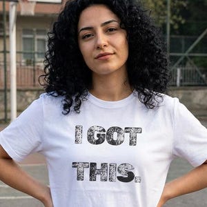 Puede incluir: Camiseta blanca con el texto "I GOT THIS" en negro, con textura. La camiseta es de manga corta y la lleva una persona con pelo oscuro y rizado. La persona está de pie al aire libre en una cancha de baloncesto.