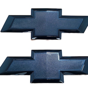 May include: Two black Chevrolet bow tie emblems. The emblems have a glossy black border and a textured, dark blue center. The design is a classic cross-like shape, commonly used as a car logo.