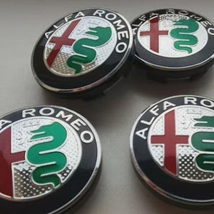 May include: Four Alfa Romeo wheel center caps. Each cap features the Alfa Romeo logo: a red cross, a green serpent, and the words "ALFA ROMEO" in a circular design. The caps have a black background and a silver border.