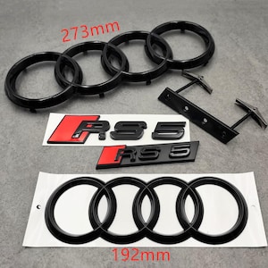 May include: Black car emblems and badges, including the Audi rings and RS5 logos. The Audi rings measure 273mm and 192mm. The RS5 emblems have a black finish with red accents. Automotive accessories.