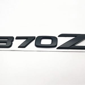 May include: Black 370Z emblem with a matte finish. The numbers and letter are in a bold, sans-serif font. The emblem is on a white background.