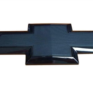 May include: A black Chevrolet emblem in the shape of a cross. The emblem has a glossy black border and a textured, dark gray center. The emblem is a car accessory.