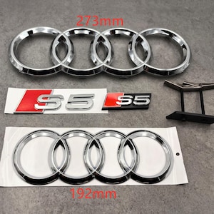 May include: Chrome car emblems, including a large set of four interlocking rings measuring 273mm, and a smaller set at 192mm. Two S5 emblems with red and silver details are also shown. A black display stand is visible.