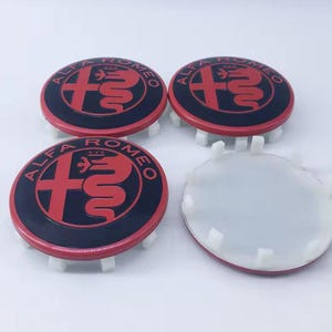 Op de afbeelding: Vier Alfa Romeo wieldopjes. De doppen zijn rond met een zwarte buitenring en een rode midden met het Alfa Romeo logo. Eén dop mist zijn midden. De doppen hebben witte plastic montage lipjes.