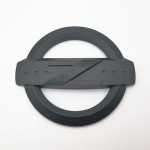 Puede incluir: Un emblema negro mate con un diseño circular y una sección central rectangular. El centro tiene una línea diagonal y pequeños recortes rectangulares. Probablemente para uso automotriz.