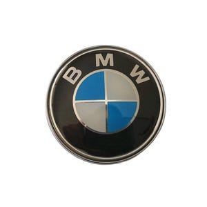 May include: A circular black emblem with a silver edge. The centre has a divided circle in blue and silver, with the letters "BMW" in white. This is likely a car badge or logo.