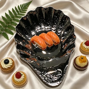 Könnte beinhalten: Ein glänzendes, silberfarbenes, muschelförmiges Serviertablett enthält drei Lachssushi-Stücke. Kleine Törtchen mit Fruchtbelägen, darunter Blaubeeren, Himbeeren und Mango, sind um das Tablett auf einer weißen Satin-Oberfläche angeordnet. Ein Farnblatt befindet sich im Hintergrund.