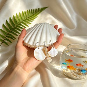 Könnte beinhalten: Ohrringe mit Perlmutt-Finish, präsentiert auf einer großen weißen Muschel. Die Ohrringe haben eine runde, rechteckige und fächerförmige Form. Ein Farnblatt und ein Fischglas mit Fischen befinden sich im Hintergrund.