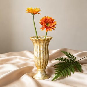 Könnte beinhalten: Eine dekorative, strukturierte, goldfarbene Vase mit zwei orangefarbenen Gerbera-Gänseblümchen und grünen Stielen. Ein grünes Farnblatt liegt auf dem drapierten, beigen Stoff. Die Vase ist etwa 15 cm hoch.