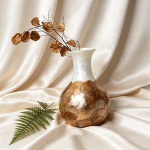 Könnte beinhalten: Eine dekorative Vase mit einem weiß schimmernden Oberteil und einem unteren Bereich, der mit sich überlappenden, irisierenden, braun getönten Muschelteilen verziert ist. Die Vase enthält getrocknete Samenkapseln auf schlanken Stielen. Ein grüner Farnwedel liegt in der Nähe.