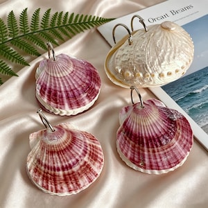 Puede incluir: Cuatro objetos decorativos en forma de concha con anillos de metal plateado. Tres son una mezcla de blanco, rosa y burdeos. El cuarto es una concha grande de color blanquecino. Los artículos están dispuestos sobre una superficie de satén con una revista que dice "Orcait Breams".