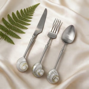 May include: A set of stainless steel cutlery including a knife, fork, and spoon. Each piece has a long handle and a decorative seashell base. The set is presented on a cream satin surface, with a green fern leaf in the background.