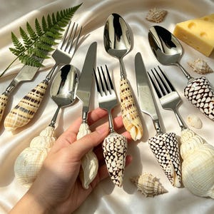 May include: A collection of stainless steel silverware with handles made from seashells. The set includes forks, knives, and spoons. The seashells are cream-colored with brown and black patterns. The silverware is arranged on a cream-colored satin surface.
