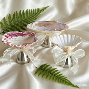 Könnte beinhalten: Drei dekorative Muschelschalen mit silbernen Sockeln und weißen Schleifen. Die Muscheln zeigen natürliche Farben von Rosa, Weiß und Braun. Ein grünes Farnblatt liegt in der Nähe der Schalen auf einer weißen Satin-Oberfläche.