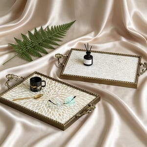 Puede incluir: Dos bandejas rectangulares con asas decorativas. Una tiene un diseño de mosaico blanco, la otra un estampado floral. Una taza negra, una cuchara y gafas están en una bandeja, y un frasco difusor en la otra.
