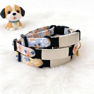 May include: Two floral patterned dog collars with black buckles and silver nameplates. One collar has a pink and orange floral pattern, and the other has a blue and orange floral pattern. A small dog figurine is in the background.