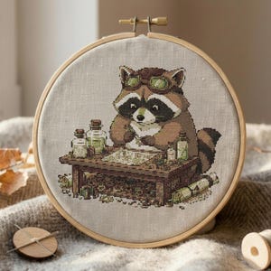 May include: Cross-stitch embroidery of a raccoon wearing goggles, seated at a table with bottles and a map. The raccoon is brown, white, and black, with a detailed design. The embroidery is set in a wooden hoop.