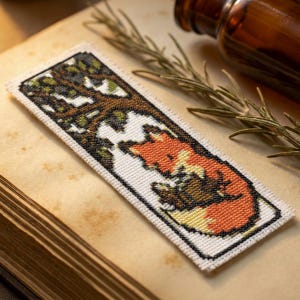 Puede incluir: Marcapáginas hecho a mano con un diseño pixelado de un zorro bajo un árbol. El zorro es naranja, marrón y amarillo, sobre un fondo blanco con un borde negro. El marcapáginas es rectangular y descansa sobre un libro antiguo.