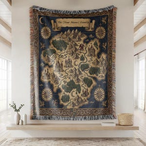 Può includere: Una coperta tessuta con un design a mappa fantasy che mostra montagne, foreste, castelli e draghi su uno sfondo blu scuro. Il banner superiore mostra il testo The [Your Name] Family. La coperta ha un bordo sfrangiato e una cornice decorativa.