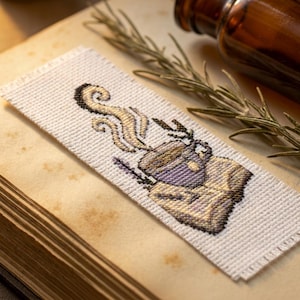 Op de afbeelding: Een rechthoekige witte stoffen boekenlegger met een kruissteekontwerp van een dampende paarse mok die op een boek rust. Het borduurwerk gebruikt paarse, gele en zwarte draden om de beker, de stoom en de details van het boek op de stof weer te geven.