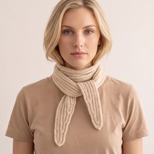 Könnte beinhalten: Ein beigefarbener Schal aus Rippenstrick, der einfach um den Hals geknotet ist und mit einem schlichten beigen T-Shirt mit kurzen Ärmeln kombiniert wird. Der Schal hat eine gleichmäßige vertikale Rippenstruktur und spitz zulaufende Enden.