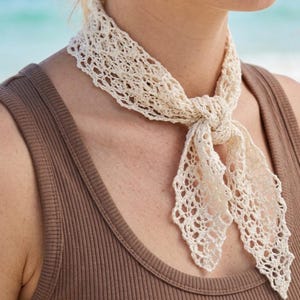 Puede incluir: Un pañuelo de cuello de ganchillo en color blanco roto con un intrincado patrón de encaje calado. El pañuelo está atado con un nudo sencillo en la parte delantera, con dos extremos puntiagudos que cuelgan. La textura delicada crea un accesorio.