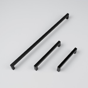 Könnte beinhalten: Drei schwarze, quadratische Schrankgriffe unterschiedlicher Länge sind diagonal auf weißem Hintergrund angeordnet. Der längste Griff ist etwa 30 cm lang, der kürzeste etwa 10 cm. Die Griffe haben eine matte Oberfläche.