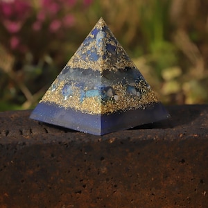 Peut inclure: Une pyramide bleue avec des paillettes et des accents dorés. La pyramide est faite d'un matériau bleu foncé et présente un design en couches. La pyramide est posée sur une surface marron foncé, avec un arrière-plan flou vert et rose.
