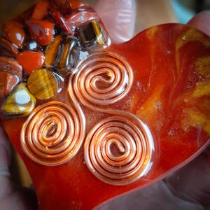 May include: A heart-shaped resin object featuring a smooth, glossy red and gold marbled surface. Embedded within the resin are three distinct copper wire spirals and a cluster of polished red jasper and golden-brown tiger's eye gemstone chips.