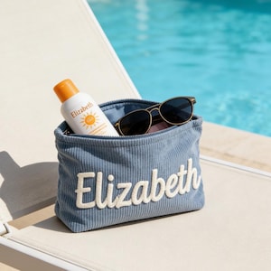 Könnte beinhalten: Eine blaue Kosmetiktasche aus Cord mit dem Namen "Elizabeth" in Weiß gestickt. Die Tasche enthält eine Sonnencreme-Flasche und eine Sonnenbrille. Das Bild wurde im Freien in der Nähe eines Pools aufgenommen.