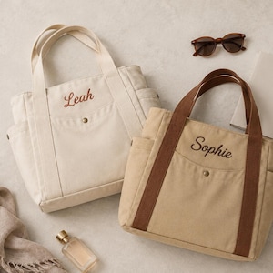 Op de afbeelding: Twee canvas draagtassen met buitenzakken en drukknoopsluitingen. De ene tas is gebroken wit met de naam Leah in rood geborduurd, de andere is beige met bruine hengsels en de naam Sophie in donkerbruin garen geborduurd.