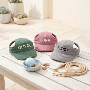 May include: Three corduroy pet hats with ear holes, personalized with the names OLIVER in green, LUNA in pink, and BUDDY in gray. A light blue tennis ball sits in a small bowl with treats, alongside a tan rope leash with gold metal hardware.