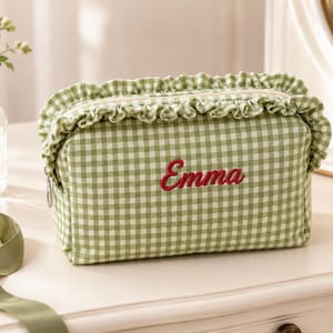 May include: A rectangular cosmetic bag featuring a sage green and white gingham pattern with a ruffled trim along the top edge. The name Emma is embroidered in red cursive script on the front centre of the bag, which includes a silver zip fastener.