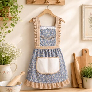May include: A blue apron with a white daisy floral pattern, featuring a white front pocket, tan polka-dot ruffled straps, and a bottom ruffle. The word Marigold is embroidered in white script on the chest, accented by white lace trim.