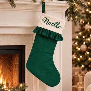 Könnte beinhalten: Ein dunkelgrüner Weihnachtsstrumpf mit einem cremefarbenen Umschlag, auf dem der Name Noelle in dunkelgrüner Schreibschrift gestickt ist. Der Strumpf verfügt über einen Rüschenbesatz unter dem Umschlag und eine passende grüne Stoffschlaufe.