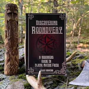May include: A book titled "Discovering Rodnovery: A Beginners Guide to Slavic Native Faith" by Perun Mountain. The book cover features a red and black design with a symbol in the center. The book is sitting on a wooden surface with a carved wooden figure and an axe.