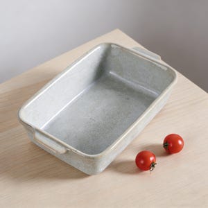 May include: A rectangular, light gray ceramic baking dish with handles. Two small red tomatoes sit beside the dish on a light wood surface. The dish has a speckled texture and a slightly glossy finish.