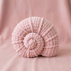 May include: A light pink, crocheted seashell-shaped cushion. The cushion features a spiral design and is made from a chunky, textured yarn. The background is a matching pink fabric, creating a cohesive and calming aesthetic.