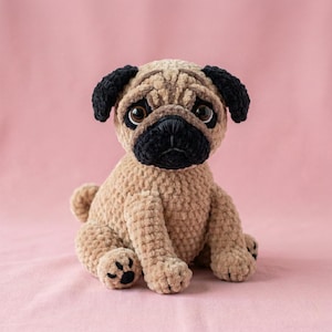 May include: A handmade plush pug dog with a tan body, black ears, and a black snout. The pug is sitting upright with large, expressive eyes. The plush toy is made of soft, textured material and is set against a pink background.