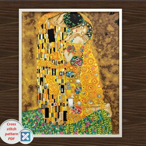 Könnte beinhalten: Ein gerahmtes Kreuzstichmuster von Gustav Klimts "Der Kuss". Das Kunstwerk zeigt zwei Figuren, die sich umarmen, mit komplizierten Mustern in Gold, Gelb und Schwarz. Der Hintergrund ist ein Mosaik aus Gold und Braun. Der Rahmen ist weiß.