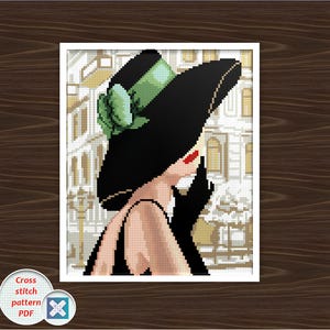 May include: Cross-stitch pattern PDF of a woman in a black hat and gloves, with red lipstick, in front of a building. The hat has a green ribbon. The image is in a white frame on a wooden background.