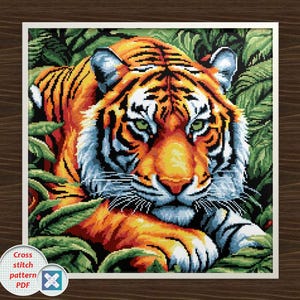 May include: A cross-stitch pattern featuring a detailed tiger. The tiger has orange and black stripes, white fur, and green eyes. It is surrounded by green foliage. The image is framed with a white border and includes the text "Cross stitch pattern PDF".