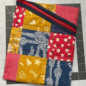 May include: A patchwork zippered pouch with a red zipper and navy blue trim. The pouch is made of various fabric squares in red with white daisies, mustard yellow with white graphics, and navy blue with white rope designs. The pouch is on a gray grid surface.