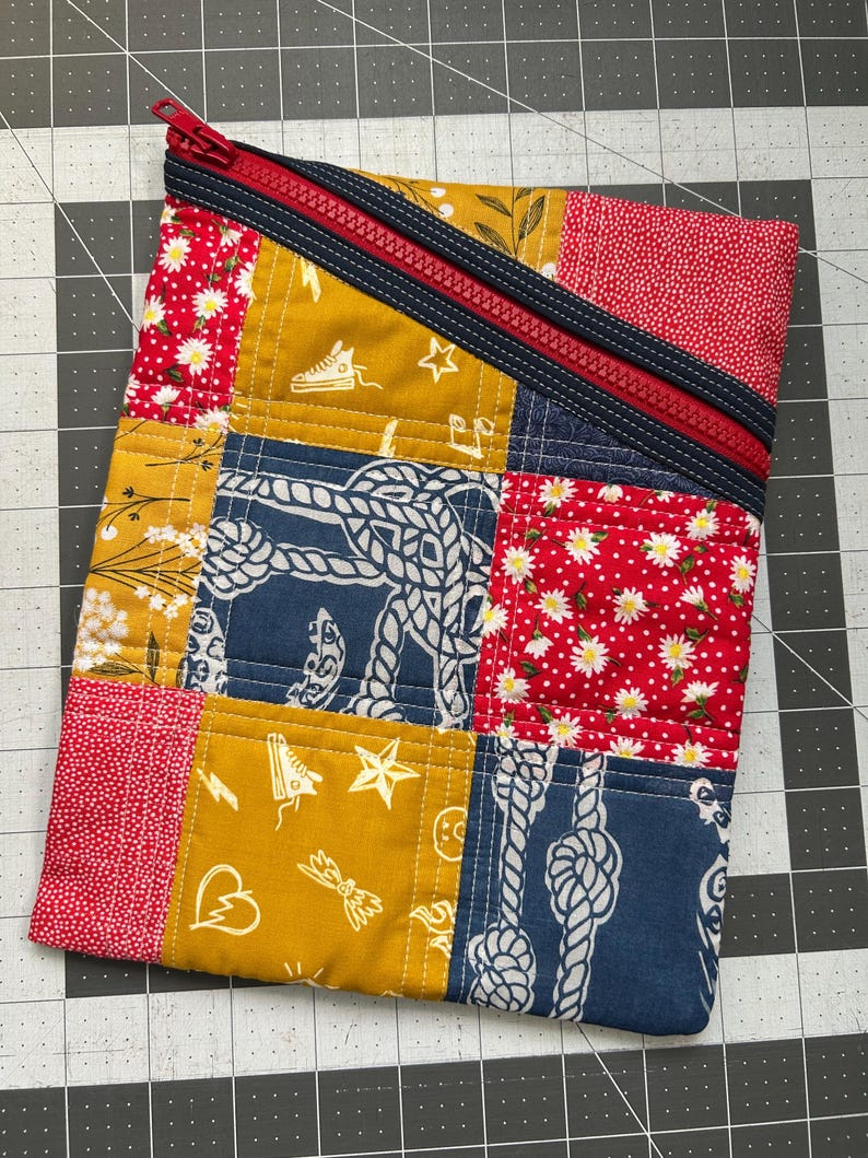 May include: A patchwork zippered pouch with a red zipper and navy blue trim. The pouch is made of various fabric squares in red with white daisies, mustard yellow with white graphics, and navy blue with white rope designs. The pouch is on a gray grid surface.