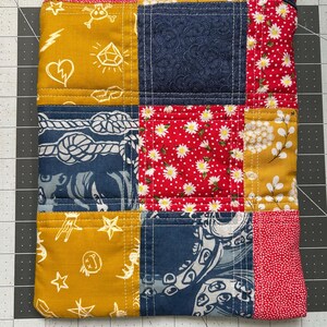 May include: A patchwork zippered pouch featuring a variety of fabrics in mustard yellow, navy blue, and red. The fabrics include floral, nautical, and patterned designs. The pouch is quilted with white thread and has a black zipper.