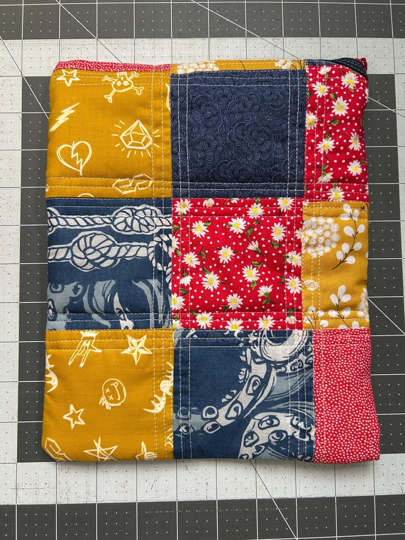 May include: A patchwork zippered pouch featuring a variety of fabrics in mustard yellow, navy blue, and red. The fabrics include floral, nautical, and patterned designs. The pouch is quilted with white thread and has a black zipper.