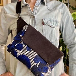 May include: A crossbody bag with a dark brown flap and strap. The bag's body features a blue floral pattern over white stripes. The bag is worn across the body, showcasing its design and functionality. The bag is a stylish accessory.
