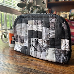 May include: A zippered pouch constructed from a patchwork of quilted fabric squares in various shades of black, gray, and white. The fabric features a mix of solid colors, floral patterns, striped textures, and abstract marbled designs.