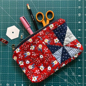 May include: A quilted fabric pouch with a red floral pattern and a navy blue zipper. The pouch is filled with sewing tools, including scissors, a pencil, and a pink pen. A white hexagon and metal clips are also visible.