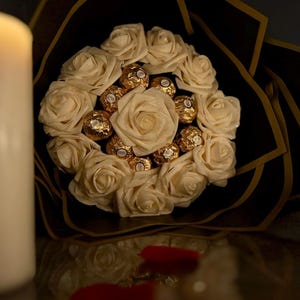 May include: A bouquet of cream-colored roses and gold-wrapped chocolates. The arrangement is set against a black background with gold accents. Red rose petals are scattered near the bouquet. A white candle is in the background.